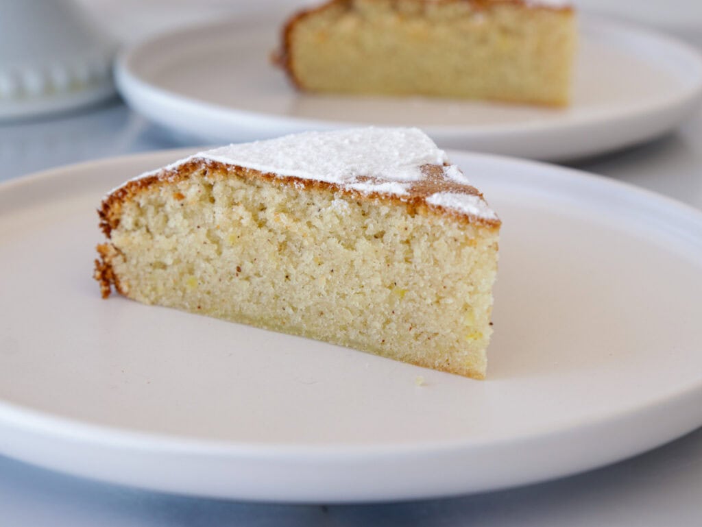 A close-up of a slice of Spanish almond cake (tarta de Santiago) on a white plate. The cake has a light, fluffy texture with a slightly browned edge and is topped with a thin layer of powdered sugar. Another slice of cake is blurred in the background on a similar plate.