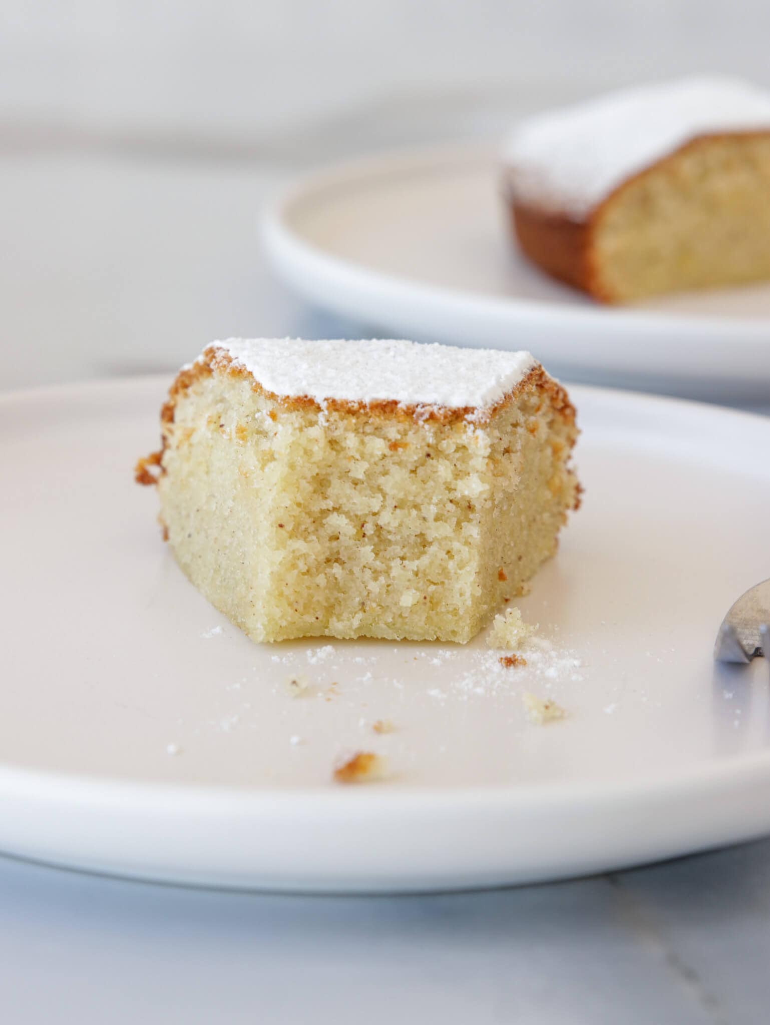 A single slice of cake dusted with powdered sugar is placed on a white plate. The cake is light and fluffy with a golden-brown crust, with a small fork bite taken out of it. Another piece of cake is blurred in the background on a similar white plate.