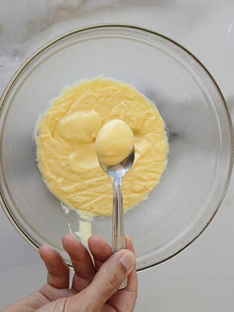 A hand holds a spoon above a glass bowl containing a creamy lemon curd mixture, likely custard or pudding, on a light-colored surface. The mixture in the bowl is smooth with some air pockets visible, indicating it has been whipped or mixed.