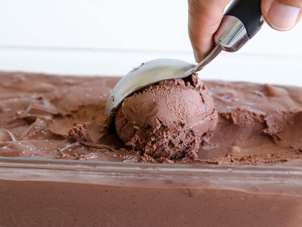 A close-up image of chocolate ice cream being scooped from a rectangular dish with a metal ice cream scooper. The rich, creamy texture of the chocolate ice cream is visible, with a hand holding the scooper.