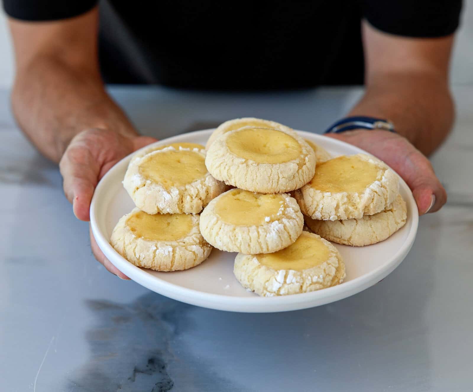 A person holds a white plate with a pile of lemon crinkle cookies. The cookies have a soft, cracked surface with powdered sugar and bright yellow centers. The person is wearing a black shirt and a watch on their left wrist.