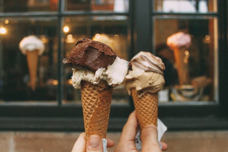 Two hands holding ice cream cones with multiple scoops of different flavors in front of a shop window displaying ice cream cones. The background is slightly blurred, showcasing the shop ambiance with lights and reflections.