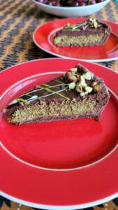 A slice of chocolate cake with a greenish filling, topped with chocolate icing, chopped nuts, and drizzled white and green icing, served on a red plate. Another slice and a bowl of fruit are in the background.
