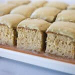 Rows of square pieces of a pistachio tres leches cake with light green frosting on top, arranged neatly on a wooden board. The cake appears soft and moist, with a fluffy texture and a thick layer of frosting.