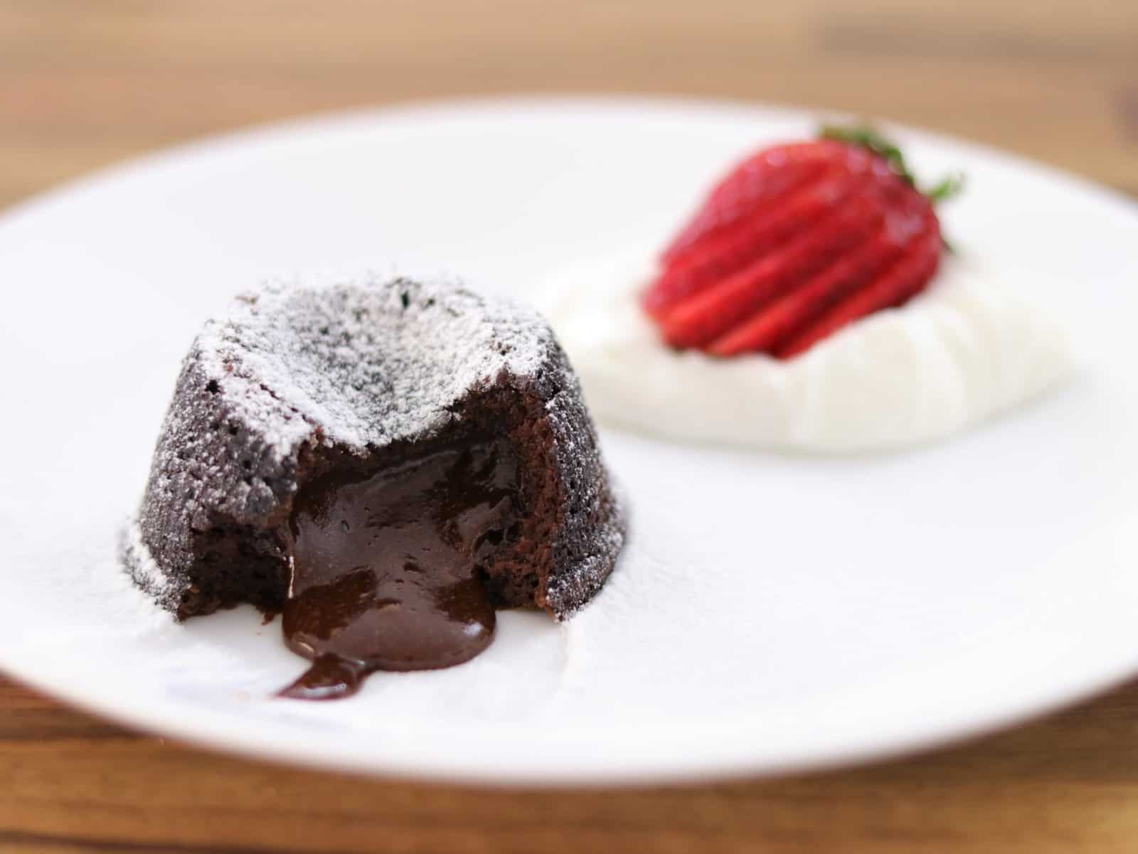 A delicious chocolate lava cake with molten center is served on a white plate. The cake is dusted with powdered sugar and is accompanied by a dollop of whipped cream topped with a sliced strawberry.