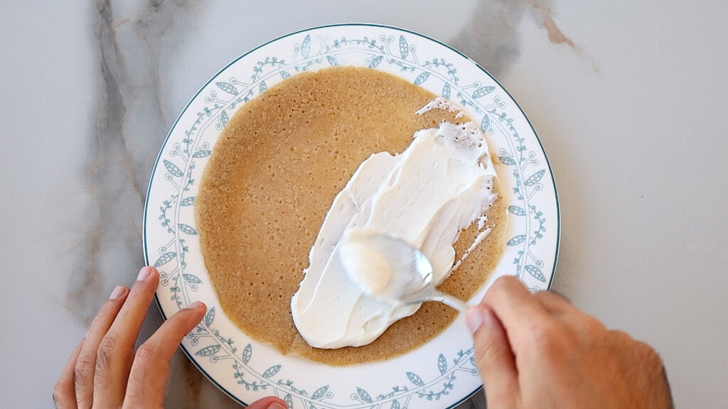 spoon spreading cream cheese on a crepe.