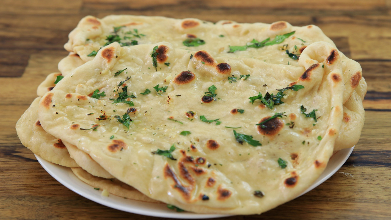 A stack of freshly baked naan bread topped with melted butter, finely chopped garlic, and sprinkled with fresh parsley. The naans are slightly golden with some charred spots, resting on a wooden surface.