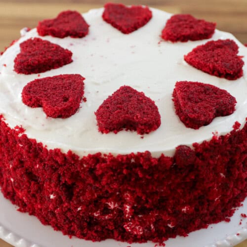 A red velvet cake with white frosting, topped with eight heart-shaped pieces of red velvet cake arranged in a circle on top. The sides are coated with red velvet cake crumbs, and the cake is placed on a white, round plate. The background is a wooden surface.