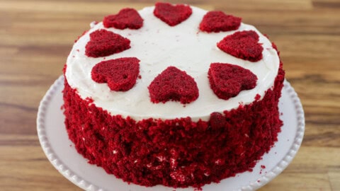 A red velvet cake with white frosting, topped with eight heart-shaped pieces of red velvet cake arranged in a circle on top. The sides are coated with red velvet cake crumbs, and the cake is placed on a white, round plate. The background is a wooden surface.