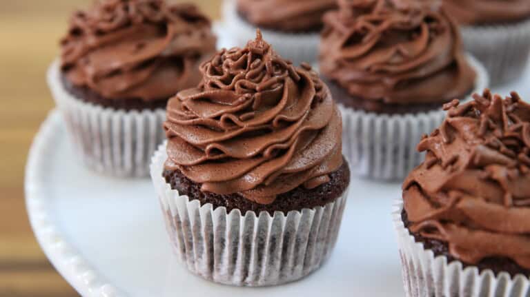 A plate of chocolate cupcakes, each topped with a generous swirl of creamy chocolate frosting, displayed in white paper liners. The cupcakes are arranged closely together in an appealing manner. The background is softly blurred, emphasizing the cupcakes.