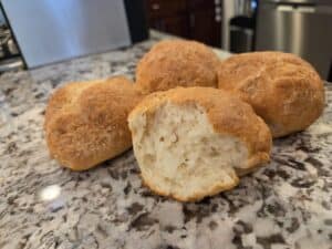 Four golden-brown bread rolls sit on a granite countertop, with one roll in the front broken open to reveal its soft, fluffy interior.