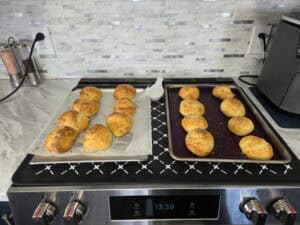 Two baking trays with freshly baked golden brown rolls topped with seasoning sit on a stovetop. The kitchen counter has salt and pepper grinders, a white spoon rest, and a modern appliance in the background.