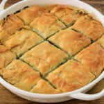 A round white baking dish filled with golden-brown, cut pieces of spanakopita, a Greek dish made with layers of flaky phyllo pastry and a spinach and feta cheese filling. The dish is placed on a wooden surface.
