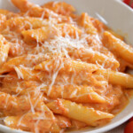 A white plate filled with penne pasta coated in a creamy tomato sauce and topped with grated cheese. Fresh cherry tomatoes on the vine are placed beside the plate.