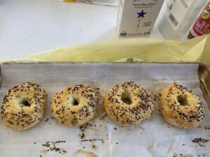 Four homemade bagels topped with black and white sesame seeds are lined up on a baking sheet with parchment paper. Baking ingredients, including sweet rice flour, are visible in the background.