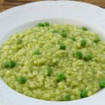 A white bowl filled with a creamy green pea risotto is placed on a wooden surface. The risotto features cooked rice mixed with bright green peas, all coated in a smooth green sauce, giving the dish a vibrant and appetizing appearance.