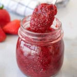 A hand holding a teaspoon of strawberry chia seed jam above a jar full of jam