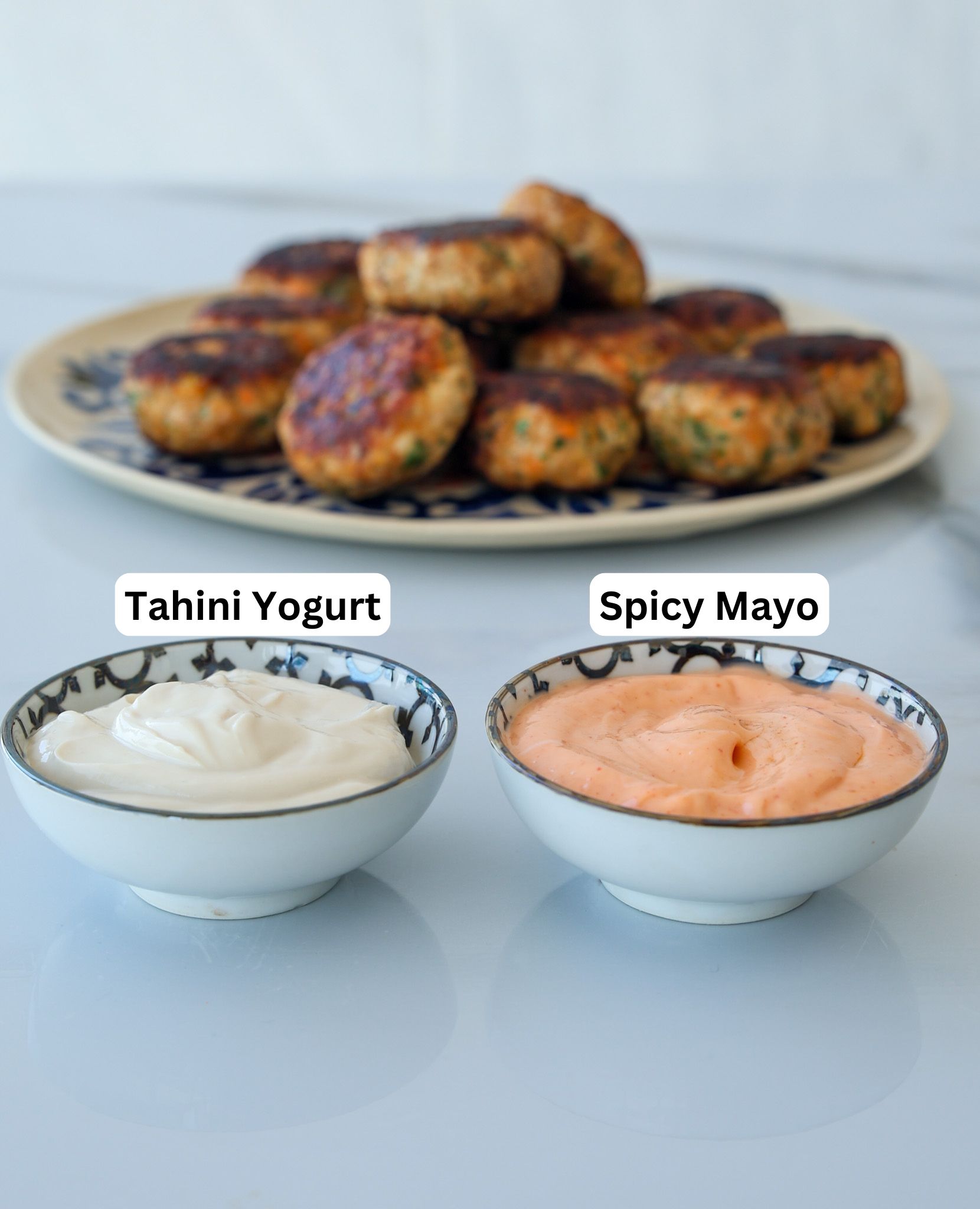 two dipping sauces on a white counder: tahini yoguet and spicy mayo. In the background there is a plate with chicken patties