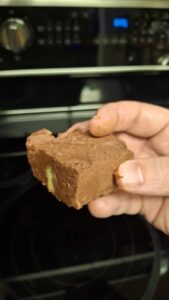A hand holds a thick, square piece of chocolate fudge with a visible walnut chunk, in front of a black oven with stainless steel dials and digital display in the background.