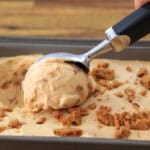 A hand uses an ice cream scoop to serve a portion of creamy, light brown ice cream topped with crumbled cookies from a metal container on a wooden surface.