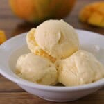 Three scoops of creamy, yellow mango ice cream in a white bowl, with chunks of fresh mango and a whole mango in the background on a wooden surface.
