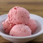 Three scoops of pink strawberry ice cream are served in a white bowl on a wooden surface.