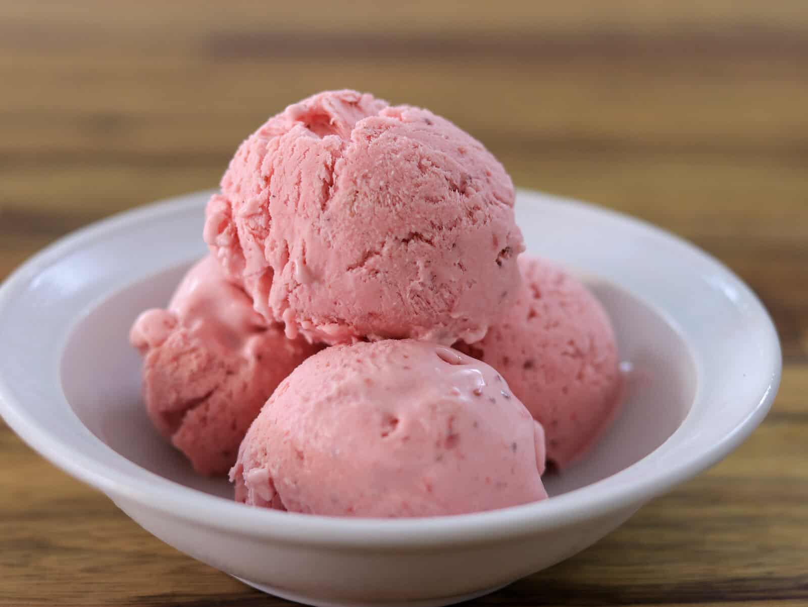 Three scoops of pink strawberry ice cream are served in a white bowl on a wooden surface.
