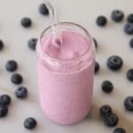 A glass of thick, pink blueberry smoothie with a clear straw is surrounded by scattered fresh blueberries on a light surface.