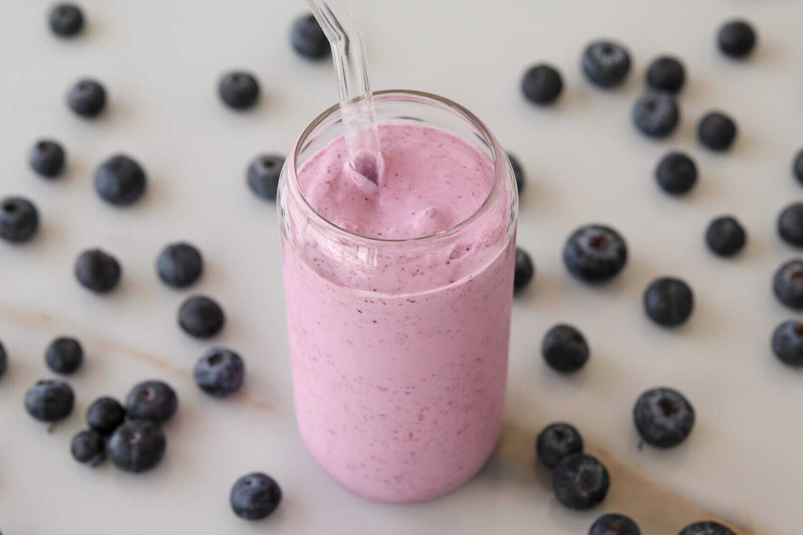 A glass of thick, pink blueberry smoothie with a clear straw is surrounded by scattered fresh blueberries on a light surface.