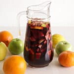 A clear glass pitcher filled with red sangria containing sliced fruits, including apples and citrus. Surrounding the pitcher are whole apples, oranges, and a lemon on a white background.