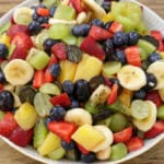 A bowl filled with a colorful fruit salad containing sliced bananas, strawberries, blueberries, grapes, pineapple, and kiwi on a wooden surface.