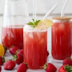 Strawberry iced tea in a glass with ice, a lemon slice, and mint garnish, surrounded by fresh strawberries and lemons. A pitcher and a jar of the same drink are in the background.