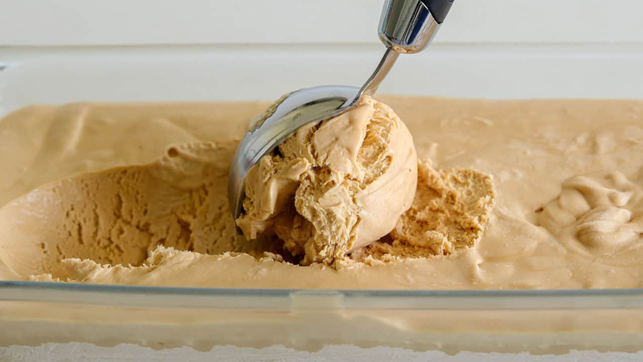 A metal ice cream scoop lifts a ball of creamy light brown salted caramel ice cream from a glass container filled with the same ice cream.
