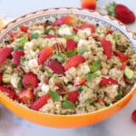 A colorful salad in an orange bowl features quinoa, fresh strawberries, avocado chunks, pecans, and mint leaves. The table is decorated with scattered strawberries, depicting a fresh and vibrant meal.