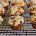 A batch of homemade blueberry muffins with crumbly streusel topping sits on a black wire cooling rack. The muffins are in colorful paper liners and appear freshly baked.