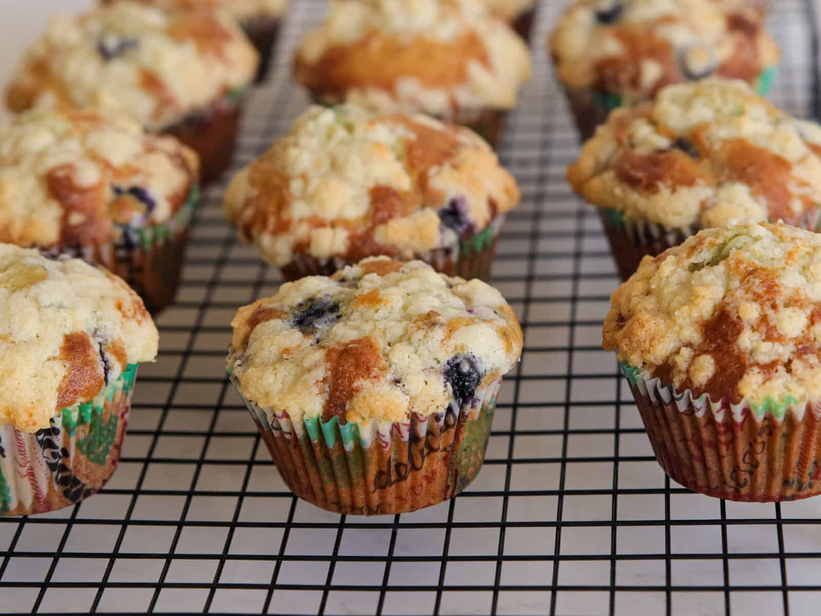 A batch of homemade blueberry muffins with crumbly streusel topping sits on a black wire cooling rack. The muffins are in colorful paper liners and appear freshly baked.