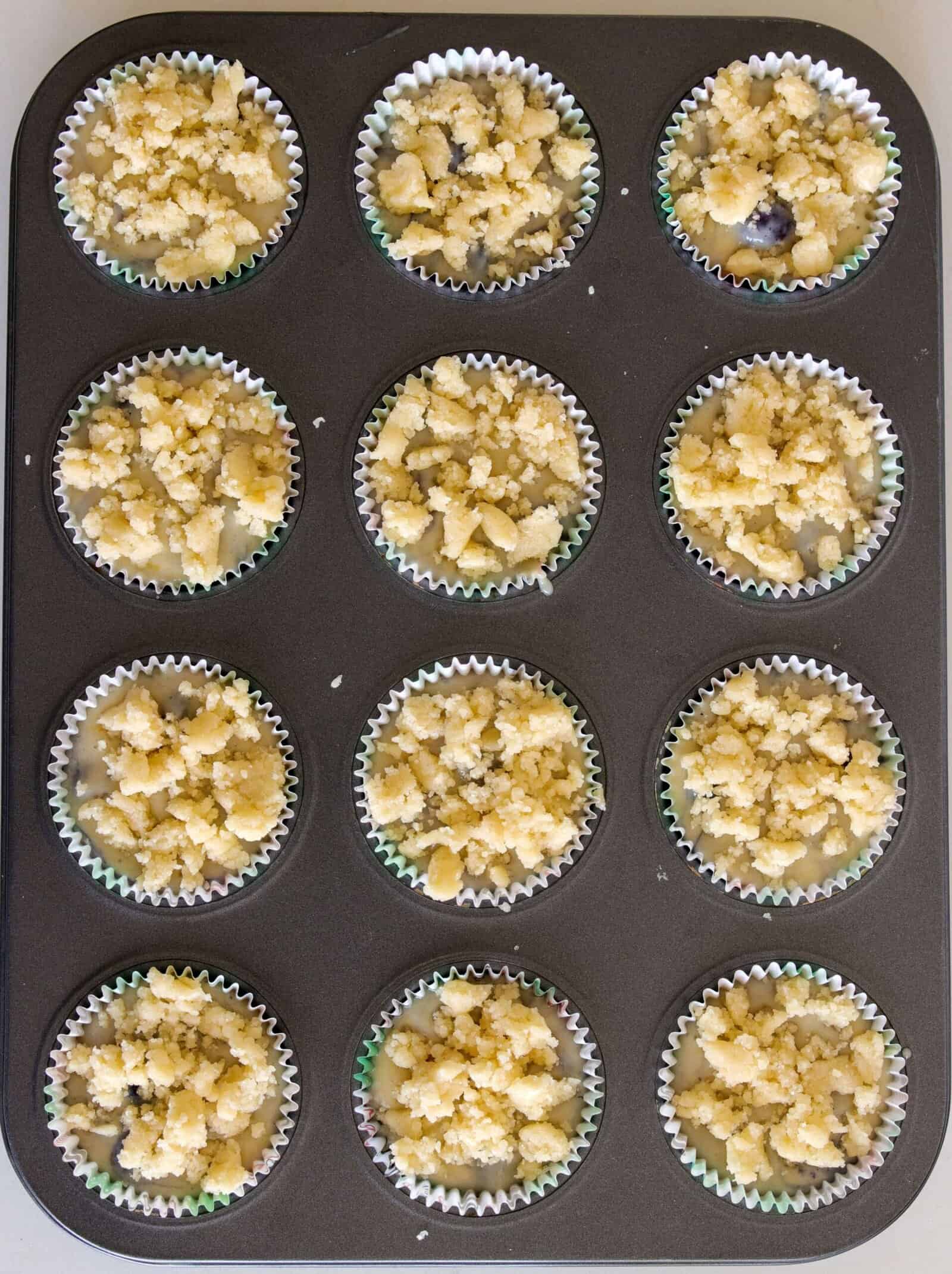 A muffin tin with twelve paper liners, each filled with unbaked crumbly streusel-topped muffin batter, viewed from above.