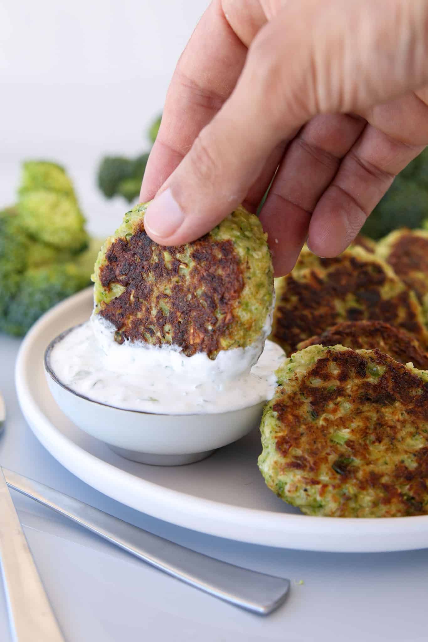 A hand dips a golden-brown broccoli fritter into a bowl of creamy white sauce, with more fritters and broccoli in the background on a white plate.