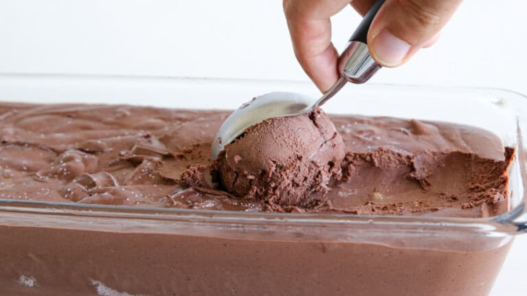 A hand uses an ice cream scoop to serve a portion of rich, creamy chocolate ice cream from a glass container.