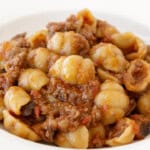 A close-up of a white plate filled with eggplant Bolognese pasta topped with a rich, chunky meat and tomato sauce. The pasta and sauce are well mixed, with visible pieces of ground meat and bits of tomato.