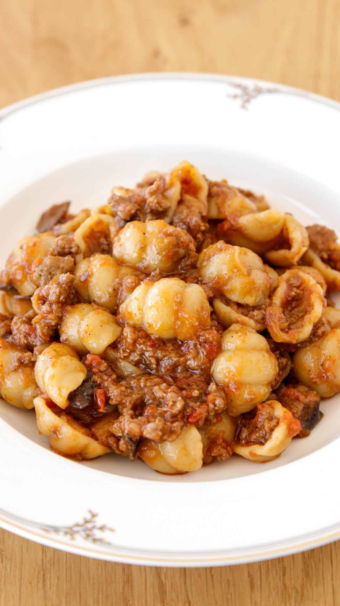 A white plate filled with orecchiette pasta mixed with a hearty tomato-based meat sauce and eggplant, placed on a light wooden surface.