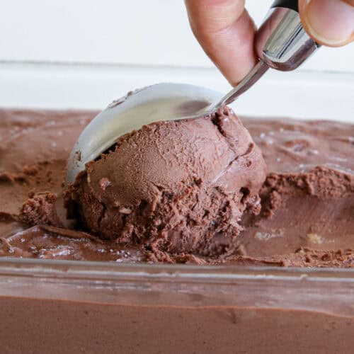 A hand uses an ice cream scoop to serve a portion of rich, creamy chocolate ice cream from a rectangular container.