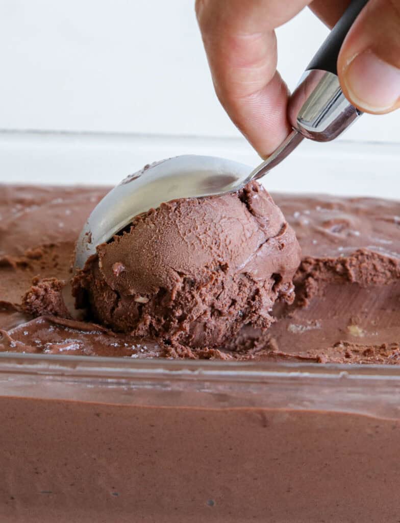 A hand uses an ice cream scoop to serve a portion of rich, creamy chocolate ice cream from a rectangular container.