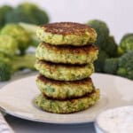 A stack of four golden-brown broccoli fritters sits on a white plate, with fresh broccoli in the background and a small bowl of sauce, a fork, and a checkered napkin nearby.