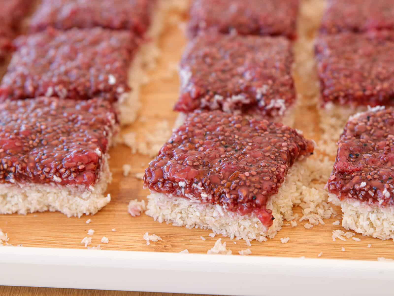 Several raspberry coconut chia seed bars arranged on a wooden tray. The bars have a white coconut base and a red raspberry chia seed topping.
