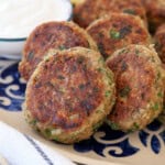 A plate of golden-brown tuna patties garnished with herbs, served with lemon wedges and a bowl of white dipping sauce on a decorative dish. A fork and napkin are nearby.