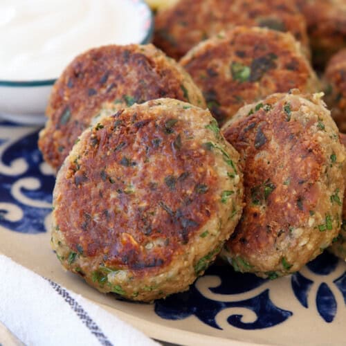 A plate of golden-brown tuna patties garnished with herbs, served with lemon wedges and a bowl of white dipping sauce on a decorative dish. A fork and napkin are nearby.