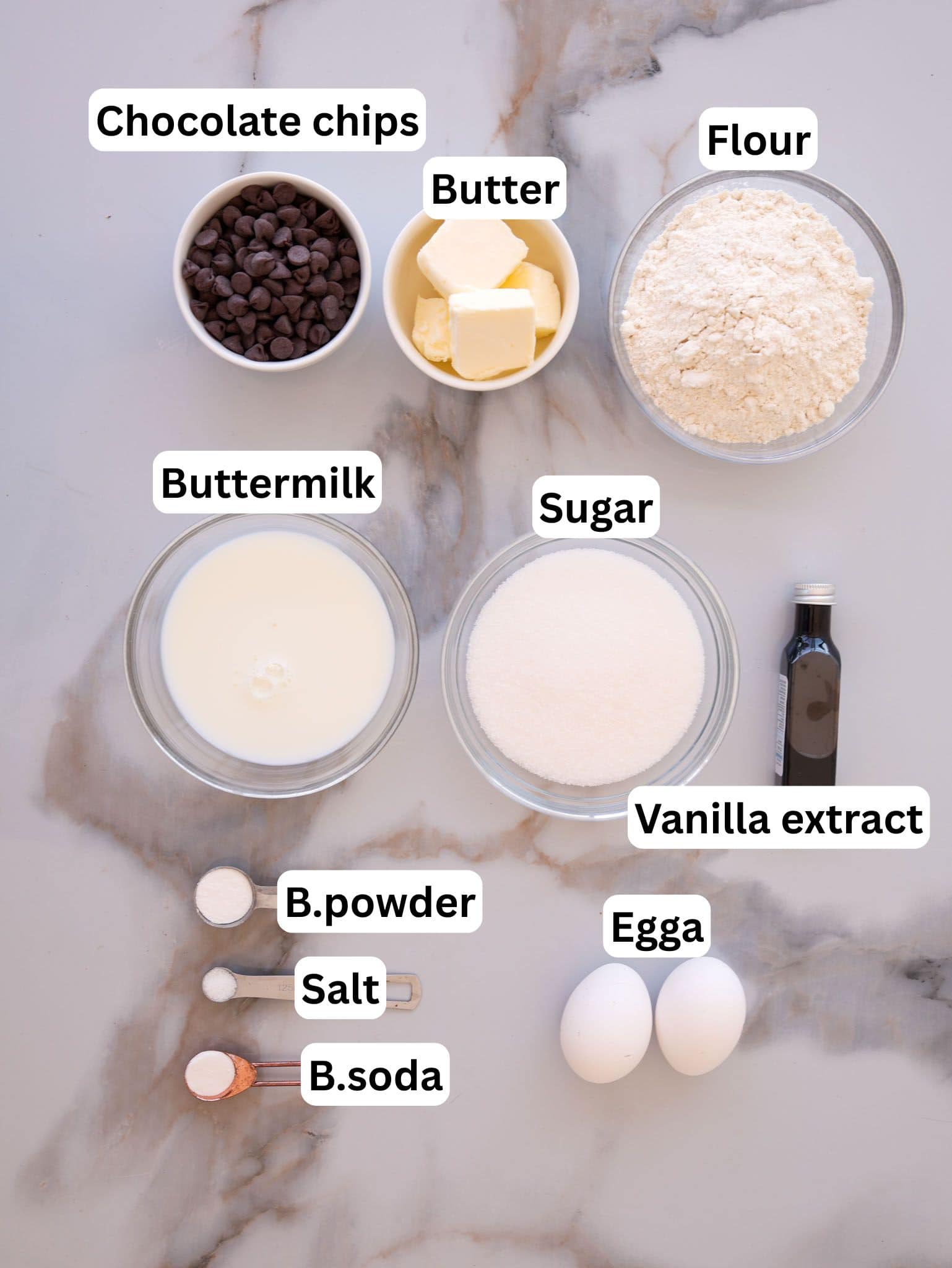Baking ingredients arranged on a marble surface: chocolate chips, butter, flour, buttermilk, sugar, vanilla extract, baking powder, salt, baking soda, and two eggs. Each item is labeled.