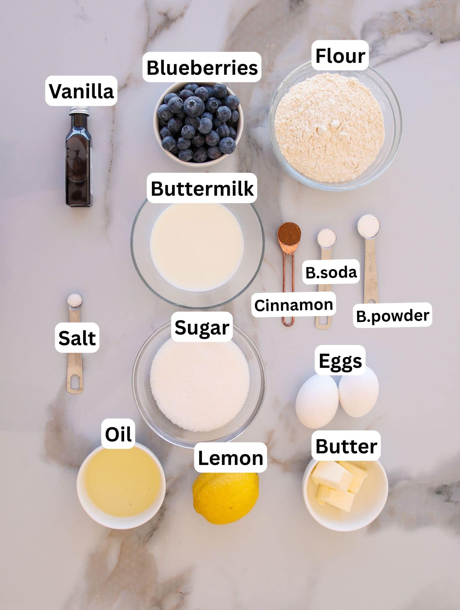 Top-down view of labeled baking ingredients on a countertop: blueberries, flour, vanilla, buttermilk, baking soda, baking powder, cinnamon, salt, sugar, eggs, butter, lemon, and oil in bowls and measuring spoons.