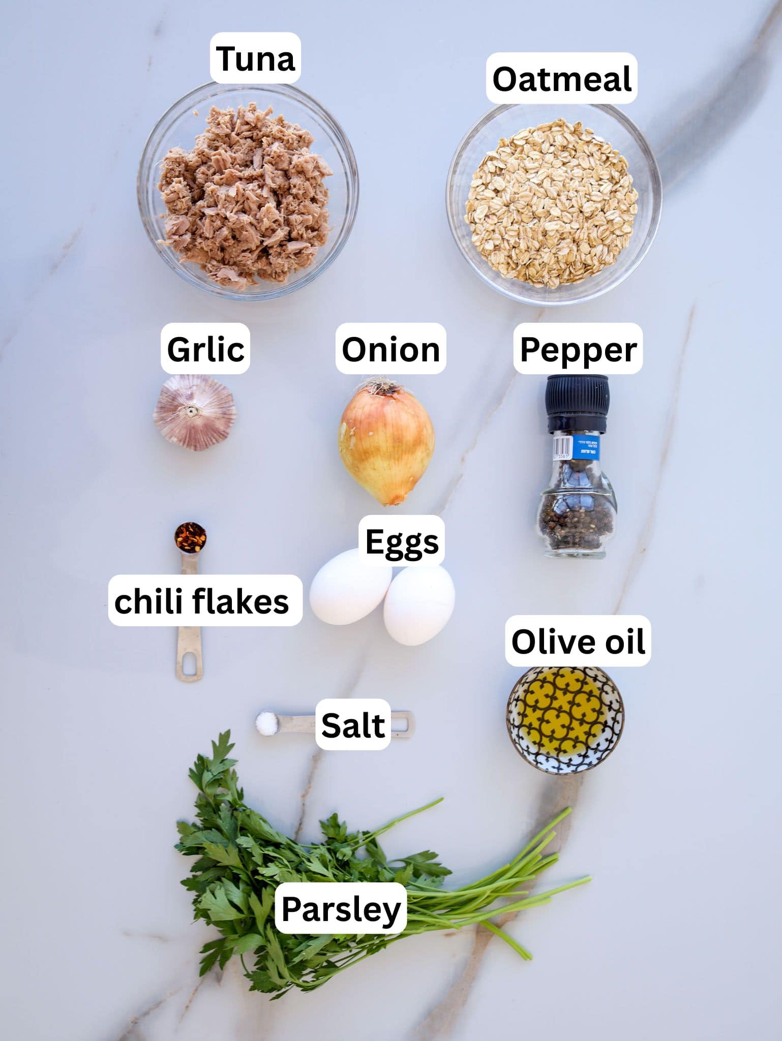 Top-down view of various ingredients arranged on a white surface, including labeled bowls of tuna, oatmeal, eggs, olive oil, garlic, onion, black pepper, chili flakes, salt, and fresh parsley.